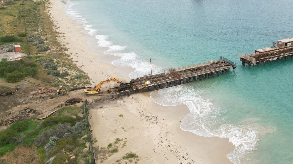 Far wide shot of the Kwinana Jetty Demolition with clear blue waters and a yellow excavator tearing apart the beach portion of the jetty.